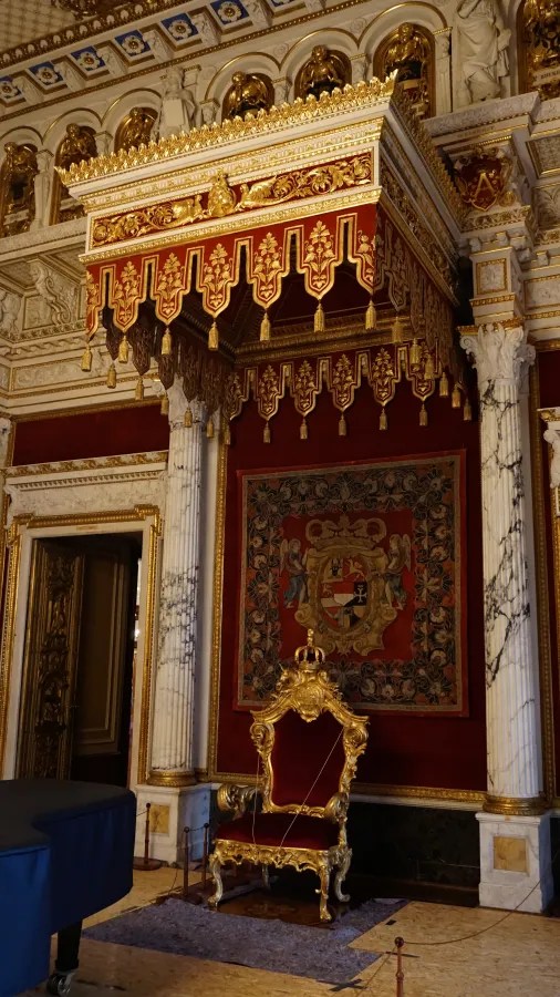 Ornate golden throne with red velvet upholstery beneath a decorative tasseled canopy in a grand hall.