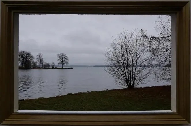 A lake view with bare trees under an overcast sky, seen through a wooden frame.