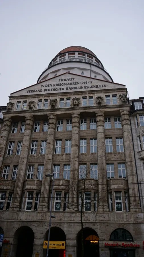 Historic stone building with columns and a dome, inscribed 'ERBAUT IN DEN KRIEGSJAHREN 1914-17 VERBAND DEUTSCHER HANDLUNGSGEHILFEN'. Banners at the base read 'WE ARE OPEN' and 'Mozart-Apotheke'.