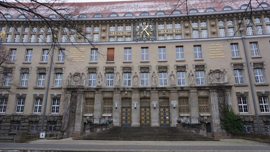 Facade of the Deutsche Bücherei building with a central clock. Visible text: KÖRPERSCHWERE LÖST DIE SCHRIFT STUMMEN GEDANKEN DURCH DER JAHRHUNDERTE STROM TRÄGT IHN ZU DIR DIE HAND; DEUTSCHE BÜCHEREI; FREIE STATT FÜR FREIES WORT FREIER FORSCHUNG SICHRER PORT REINER WAHRHEIT SCHUTZ UND HORT.