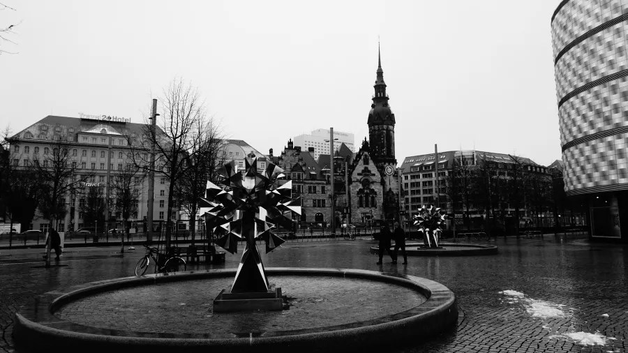 Black and white city square with geometric sculptures, a historic church tower, and Travel24 Hotel building.