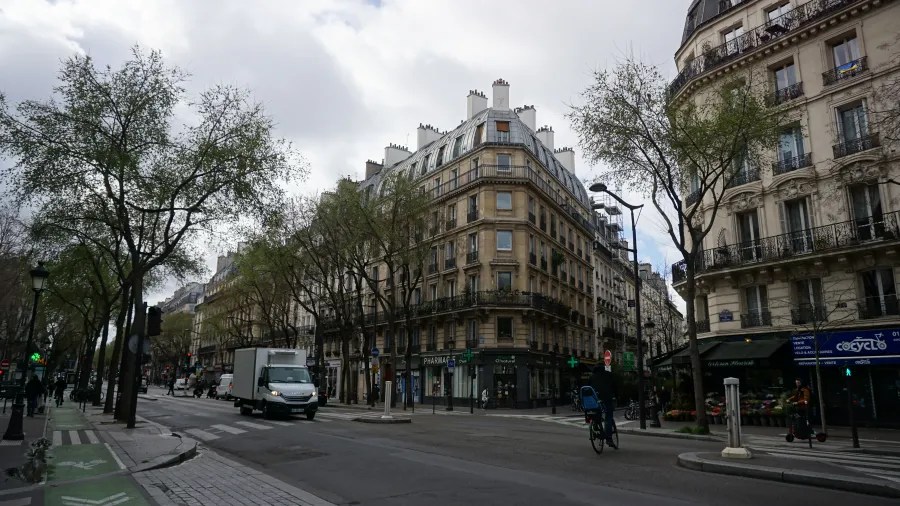 20230324_Frankreich_Paris (5) Intersection of Paris street with classic Haussmann buildings, trees lining the sidewalks, a white delivery truck, and a cyclist on the road under a cloudy sky