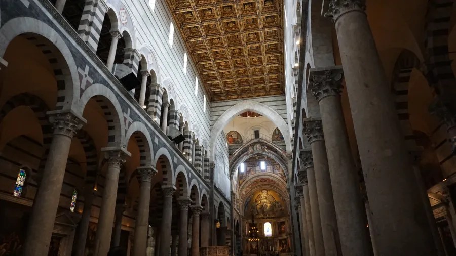 20230420_Italien_Toskana_Pisa (29) Interior view of a historic cathedral with rows of stone columns, arched windows, and detailed ceiling artwork