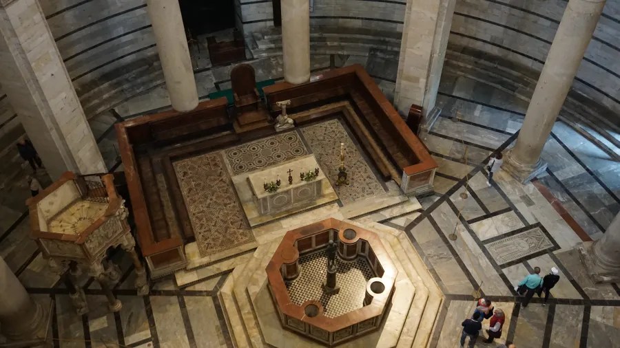 20230420_Italien_Toskana_Pisa (36) Interior view of a circular stone cathedral with a central baptismal font, wooden seating, and several visitors walking around