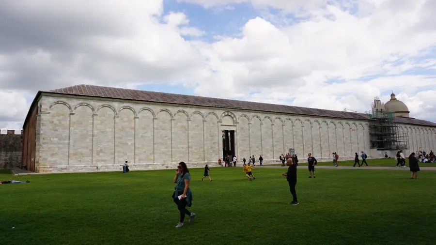 20230420_Italien_Toskana_Pisa (38) People walking and playing on green grass in front of a long white stone building with arched details and scaffolding on one side under a cloudy sky