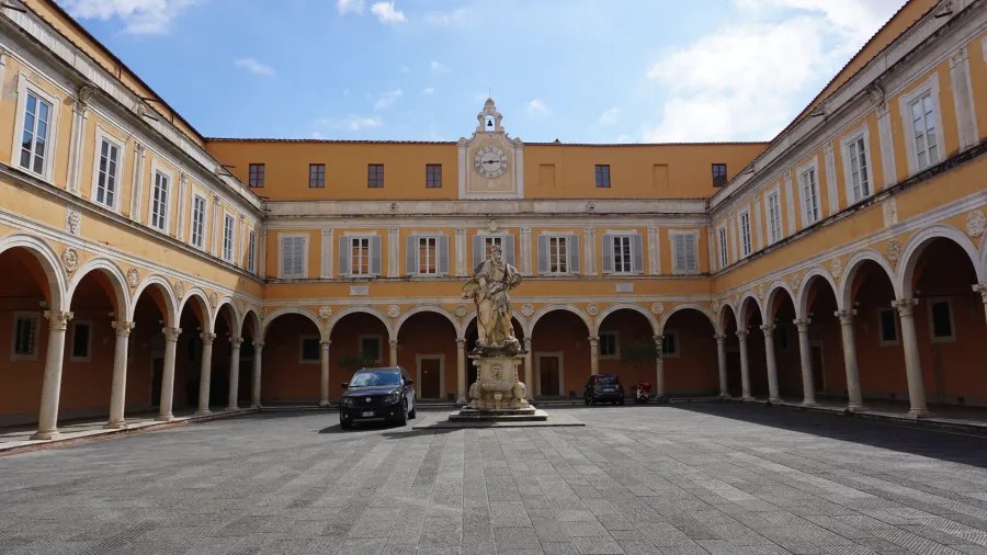 20230420_Italien_Toskana_Pisa (9) Courtyard with yellow and white arched colonnades, a central statue, and two parked cars under a partly cloudy sky