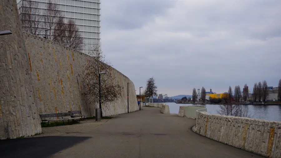 Riverside walkway with high stone walls, benches, and trees under a cloudy sky