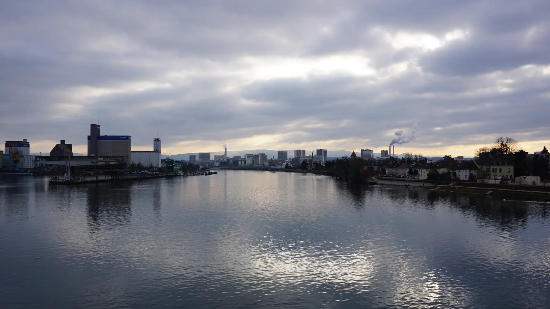 Wide river flowing through a city with industrial buildings and houses under a cloudy sky