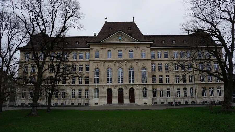Large stone building with many windows and a central clock, framed by bare trees