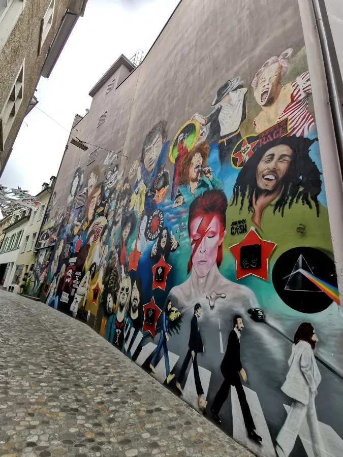 Colorful mural of iconic musicians and band logos on a narrow cobblestone street wall.