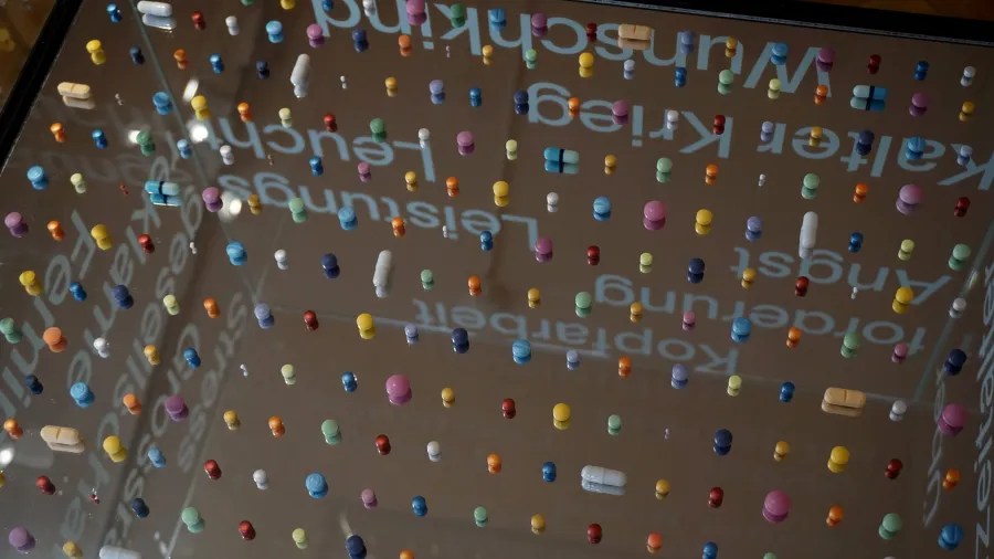 Various colorful pills and capsules arranged on a mirror reflecting German words including Wunschkind, Kalter Krieg, Angst, Kopfarbeit, Forderung, and Stress.