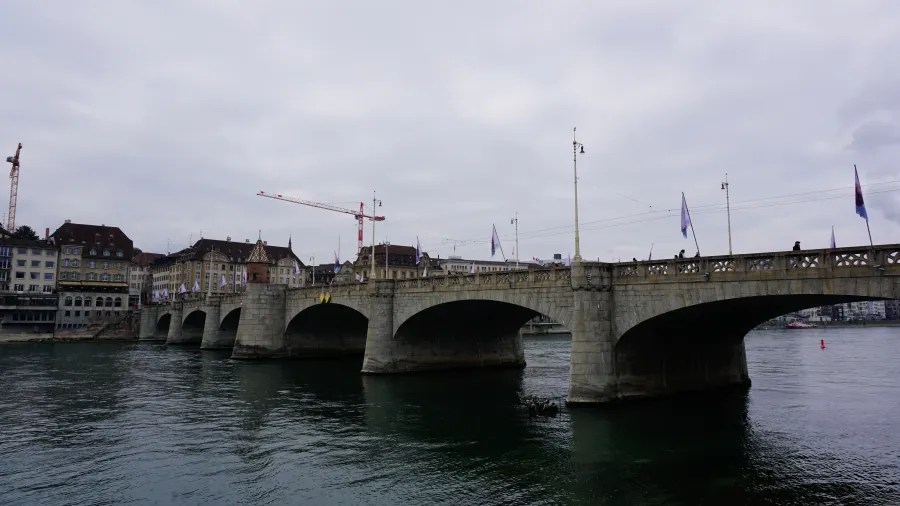 Stone arched bridge crossing a river with city buildings and cranes under an overcast sky.