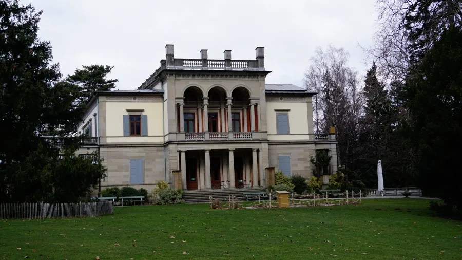 A large two-story neoclassical building with arched balconies and a green lawn in front