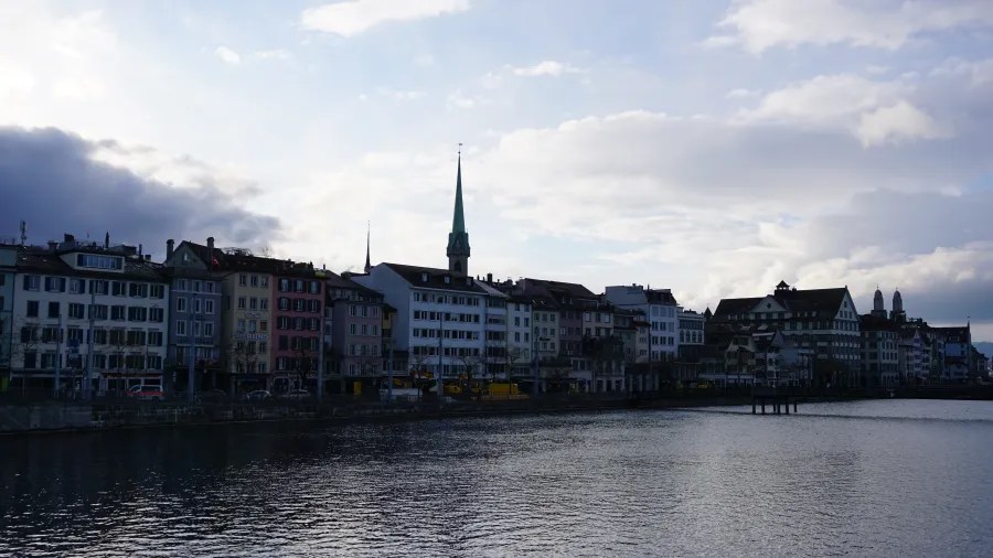 Colorful waterfront buildings and a church spire along a river under a cloudy sky.