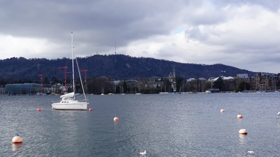 A white sailboat on a lake with a city and mountain backdrop under overcast skies.
