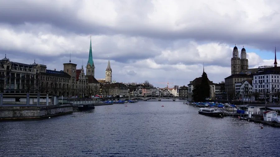 The Limmat River flowing through Zurich with prominent church spires under a cloudy sky.
