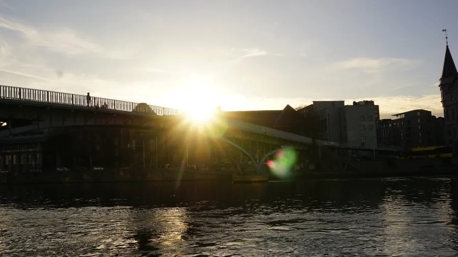 Sun shining brightly behind a bridge over a river with city buildings in the background.