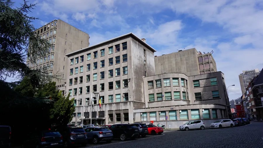 Grey concrete institutional buildings with a Belgian flag and parked cars under a blue sky