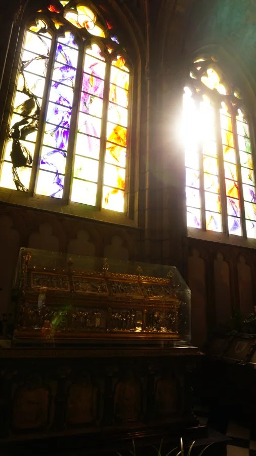 An ornate golden reliquary inside a church, positioned below sunlit stained-glass windows.