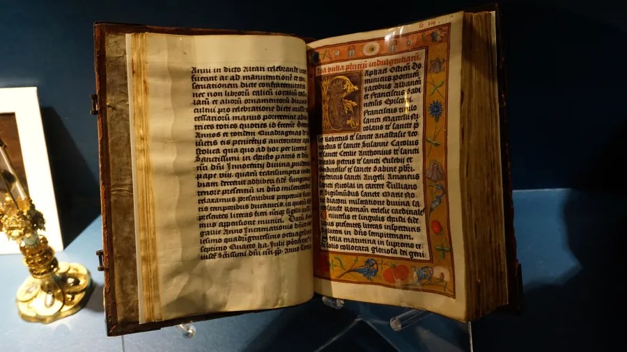 An open medieval manuscript with Latin text, an illuminated initial, and a decorative floral border.