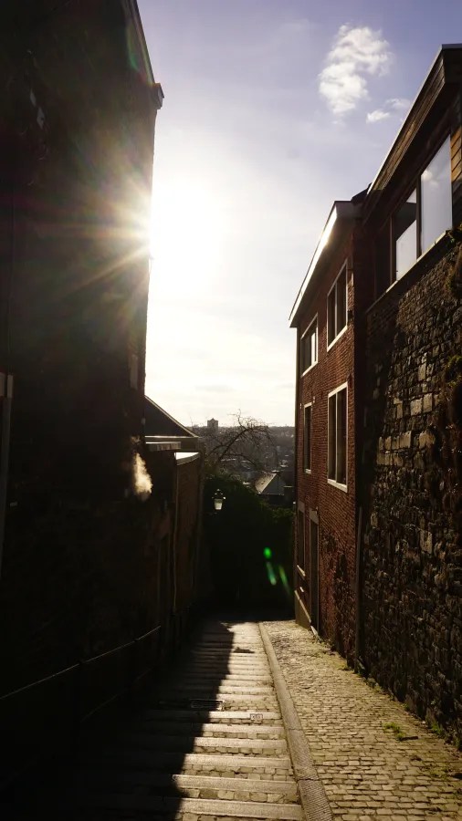 Narrow stone stairway between old brick buildings with bright sunlight creating a lens flare.