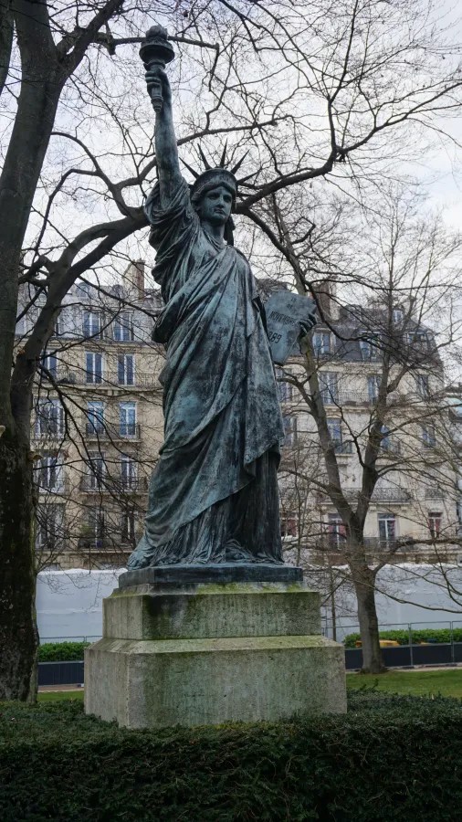20230324_Frankreich_Paris (110) Bronze statue of the Statue of Liberty holding a torch and a tablet, situated outdoors with leafless trees and multi-story buildings in the background