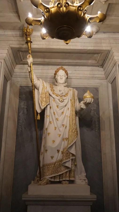 20230324_Frankreich_Paris (130) Statue of a regal figure holding a scepter and orb, dressed in ornate robes with gold details, standing inside a marble niche