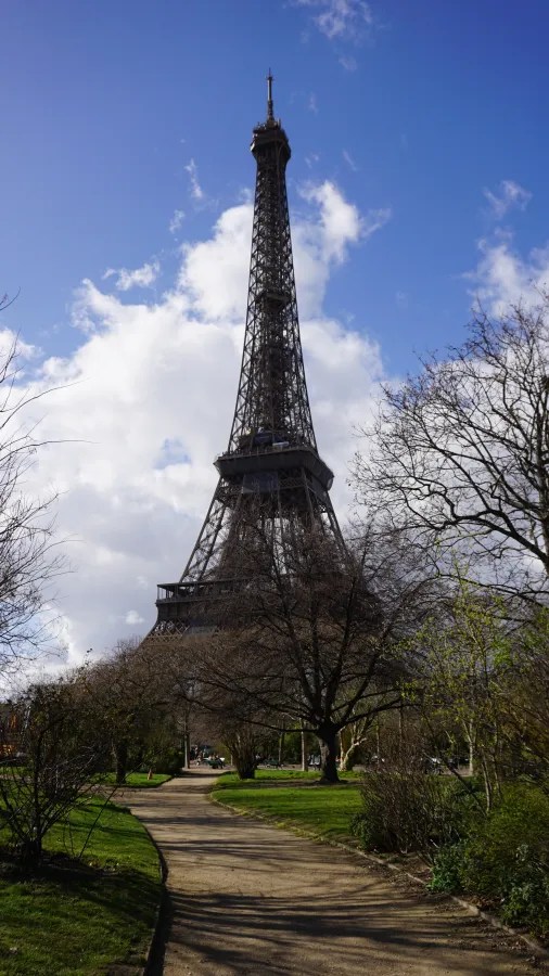 20230324_Frankreich_Paris (138) Eiffel Tower viewed from a park with a walking path and trees against a partly cloudy sky