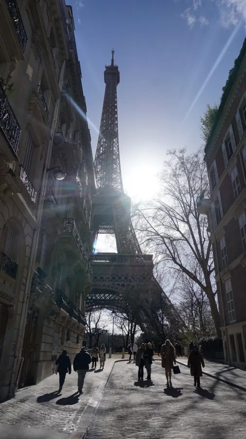 20230324_Frankreich_Paris (141) People walking near the Eiffel Tower with sun shining behind it and buildings on both sides