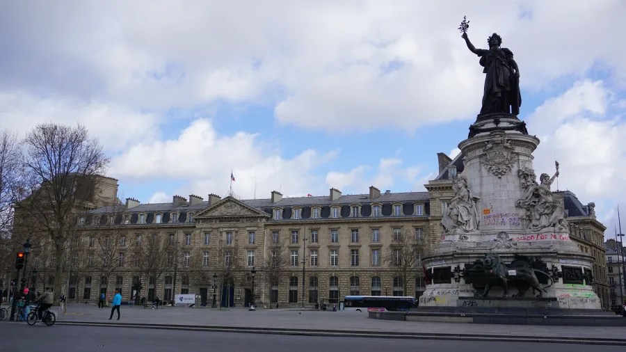 20230324_Frankreich_Paris (17) Statue of a woman holding an olive branch on a pedestal with graffiti in a public square with historical buildings in the background