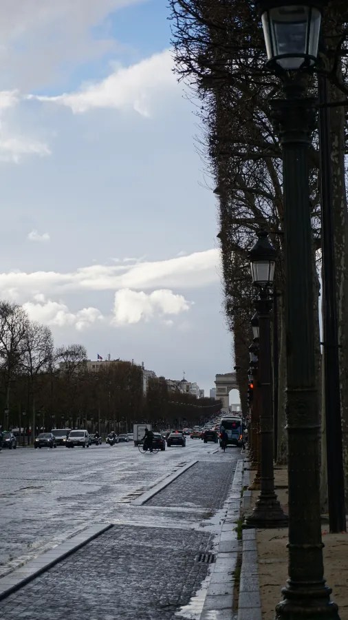 20230324_Frankreich_Paris (170) Busy city street with cars and motorcycles, lined with lampposts and trees, Arc de Triomphe visible in the distance under a partly cloudy sky