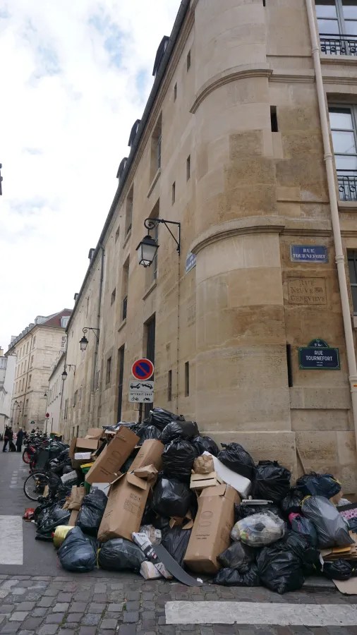 20230324_Frankreich_Paris (68) Large pile of black garbage bags and cardboard boxes stacked on a cobblestone street corner next to a beige stone building with street signs.