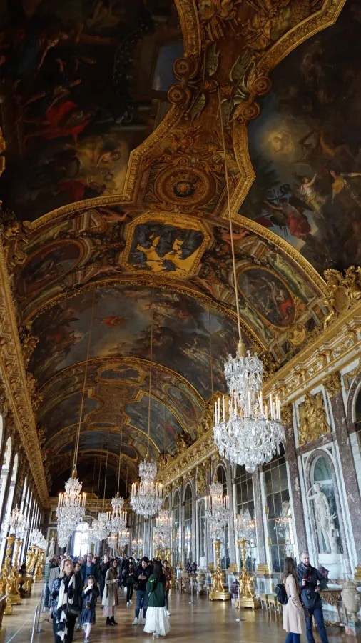 20230325_Frankreich_Paris (112) Visitors walking inside the Hall of Mirrors with ornate golden decorations, large chandeliers, and a painted ceiling