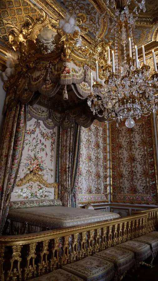 20230325_Frankreich_Paris (118) Ornate royal bedroom with floral upholstery, golden carvings, and a large crystal chandelier