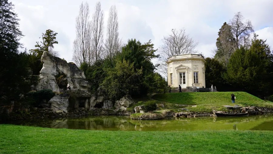20230325_Frankreich_Paris (172) Small classical stone pavilion on a grassy hill beside a reflective pond with rocky formations and trees