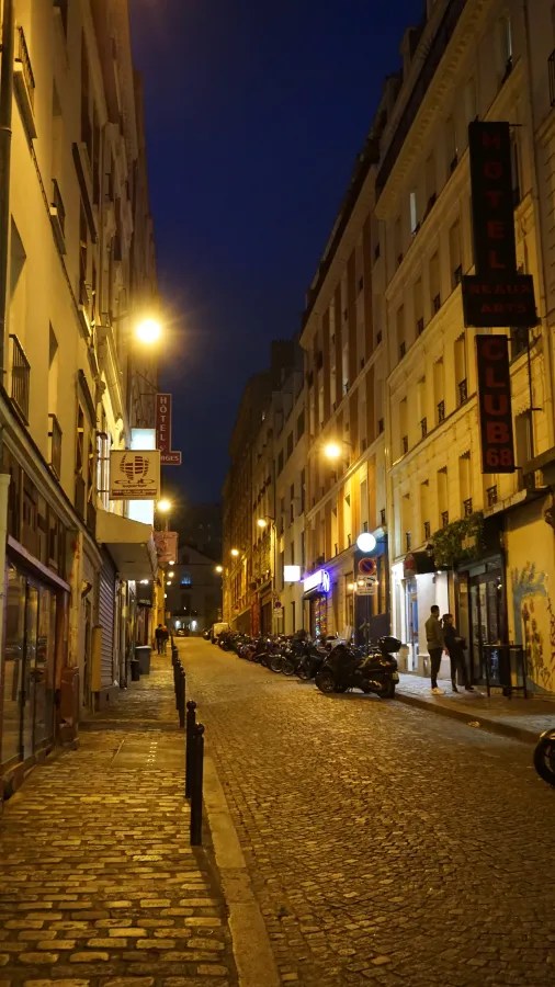 20230325_Frankreich_Paris (242) Narrow cobblestone street at night with streetlights, motorcycles parked along the side, and people walking near buildings with illuminated signs