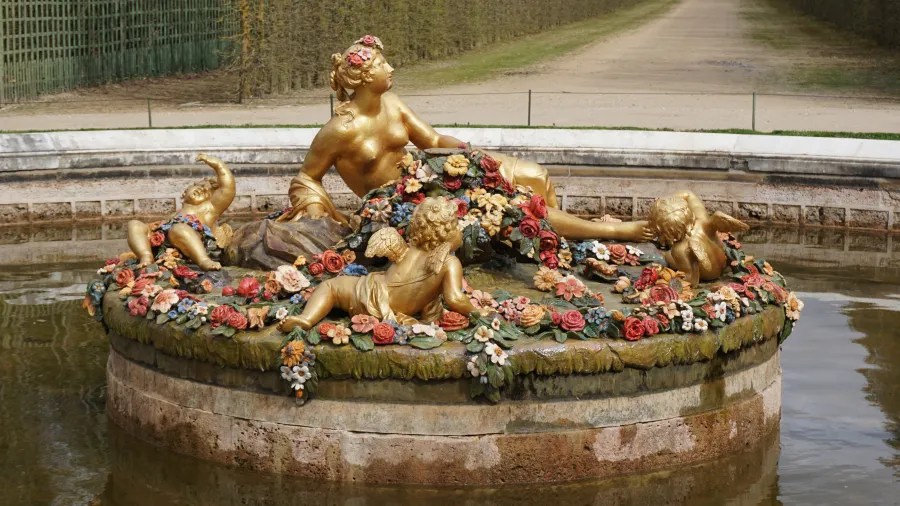 20230325_Frankreich_Paris (34) Golden statue of a reclining woman surrounded by three cherubs and colorful flowers in a circular fountain