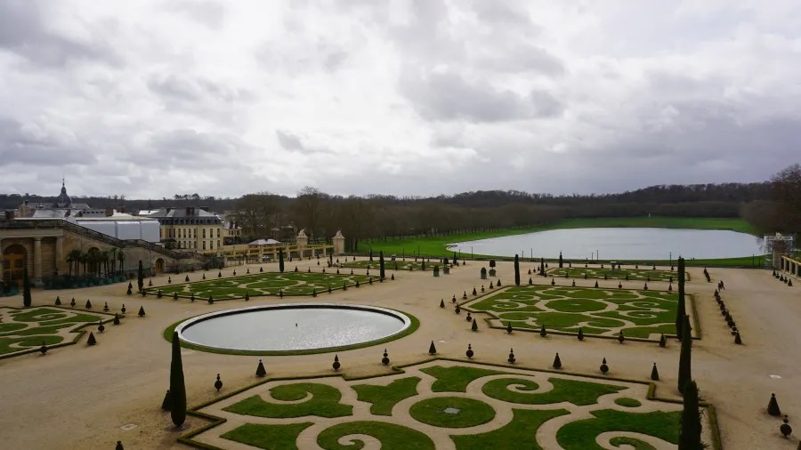 20230325_Frankreich_Paris (46) Formal gardens with symmetrical patterned hedges, gravel paths, a round reflective pond, and a large lake in the background under a cloudy sky