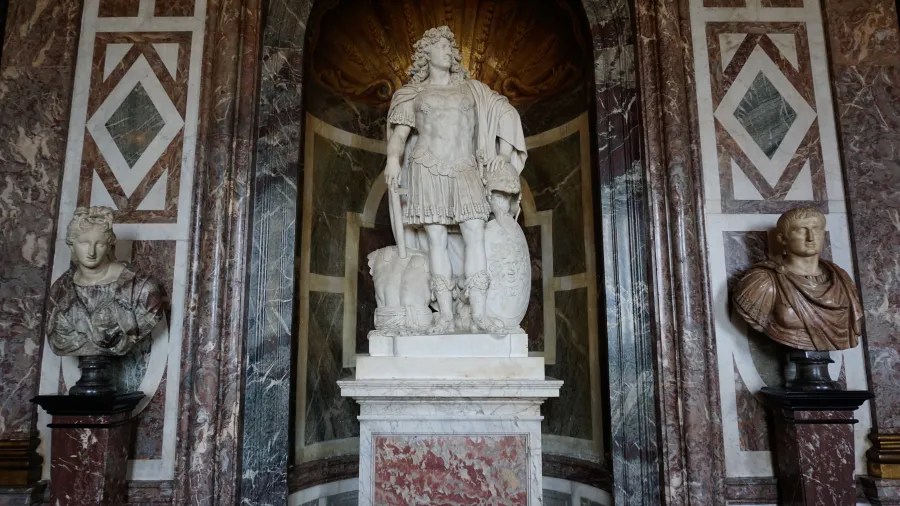 20230325_Frankreich_Paris (80) Marble statue of a Roman soldier in armor holding a shield, flanked by two marble busts inside a stone alcove with decorative patterns