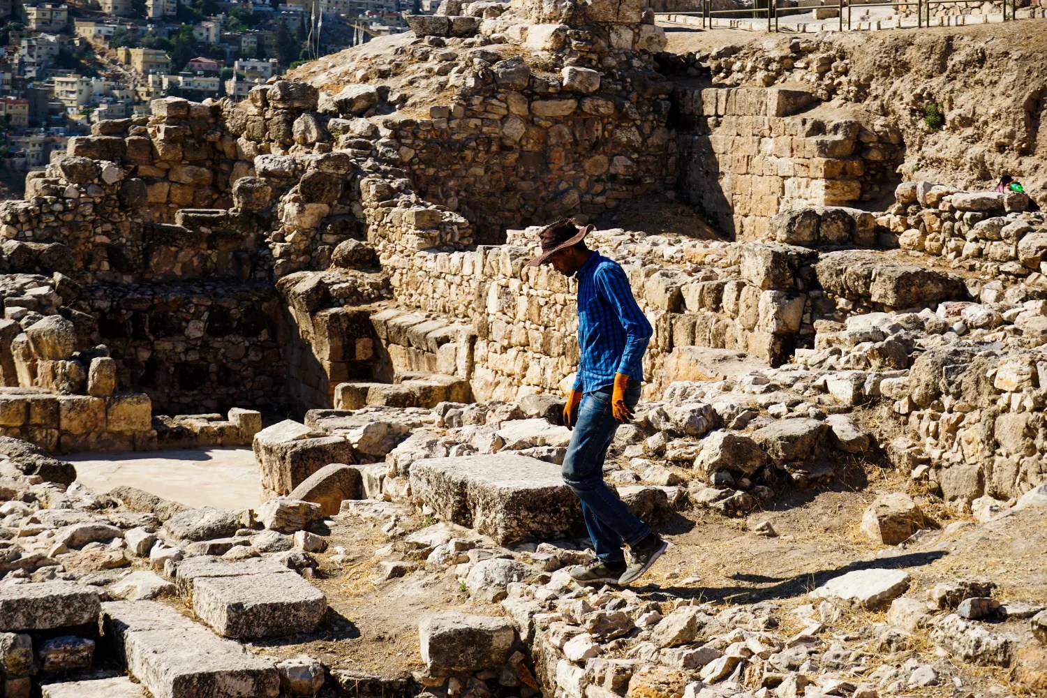 A person wearing a hat and gloves walking among ancient stone ruins in a sunny outdoor setting