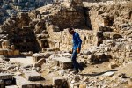 A person wearing a hat and gloves walking among ancient stone ruins in a sunny outdoor setting