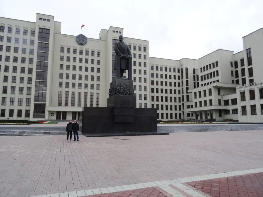 Large statue of a man on a pedestal in front of a government building with many windows and a flag on top