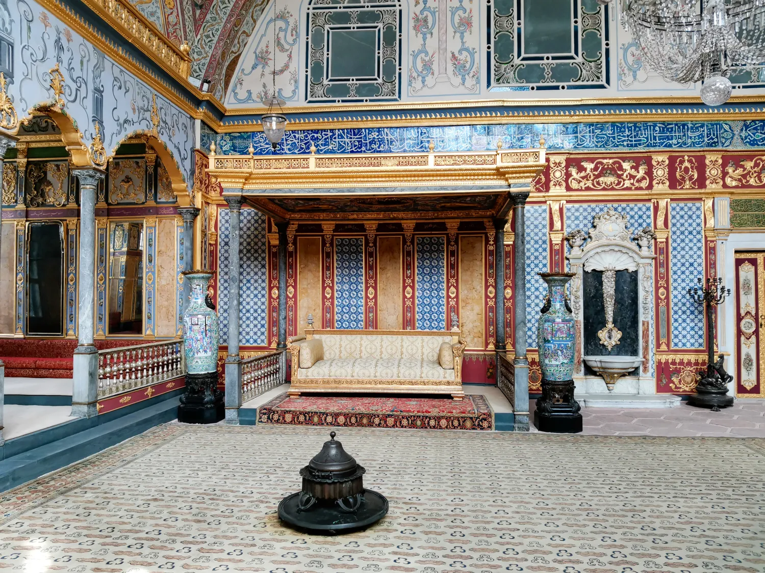 Opulent room with ornate sofa under a decorated canopy, patterned tiles, large vases, and intricate wall designs