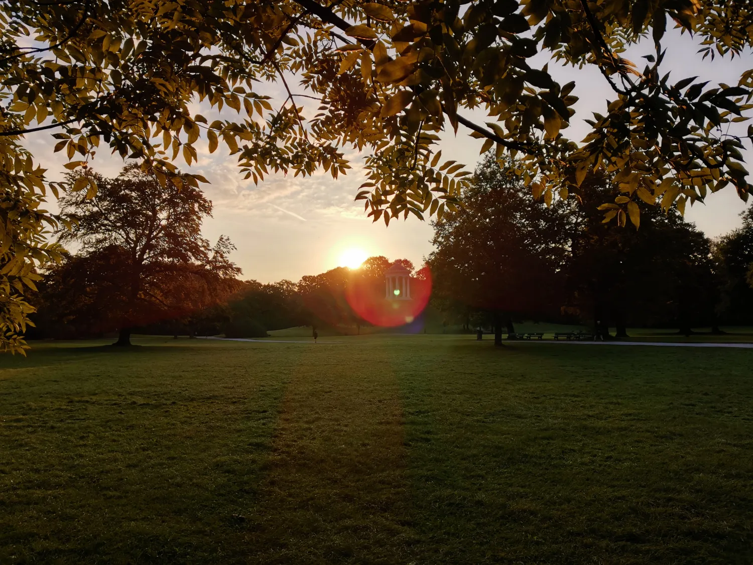 Sunset over a park with green grass, trees, and a small pavilion in the distance