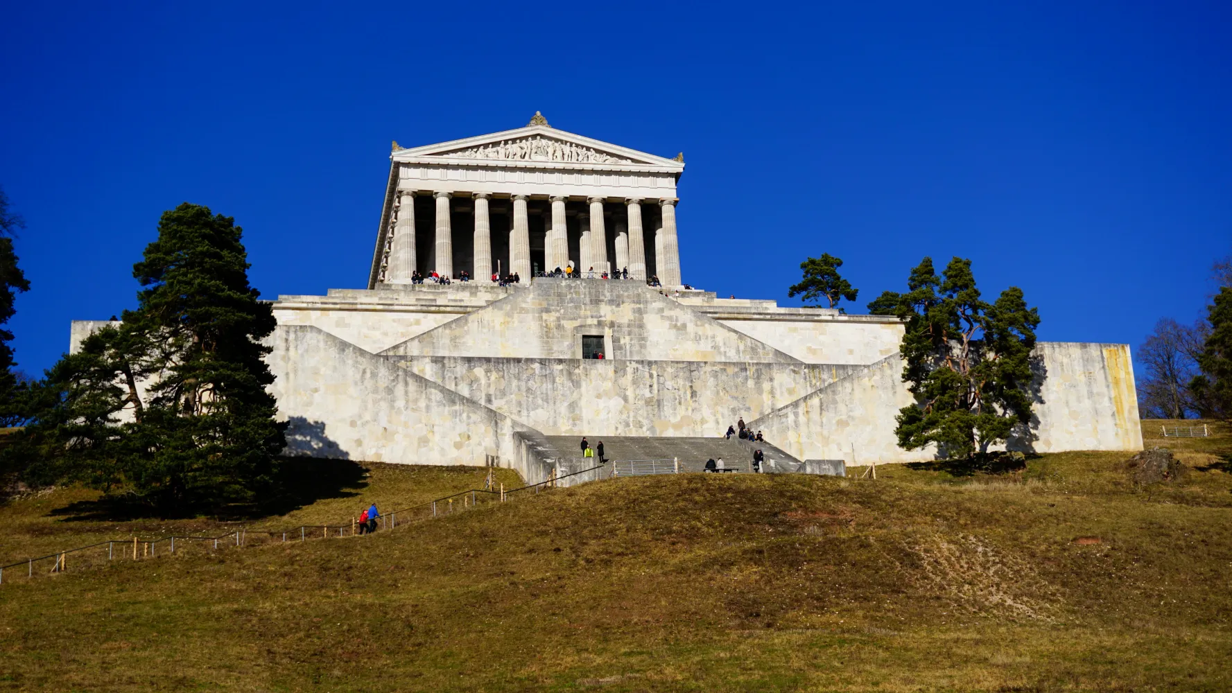A large classical monument with tall columns on a stepped stone platform against a clear blue sky, surrounded by trees and grass, with several people walking and sitting