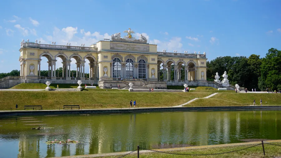 The Gloriette structure reflected in a large pond with people walking nearby and blue sky.