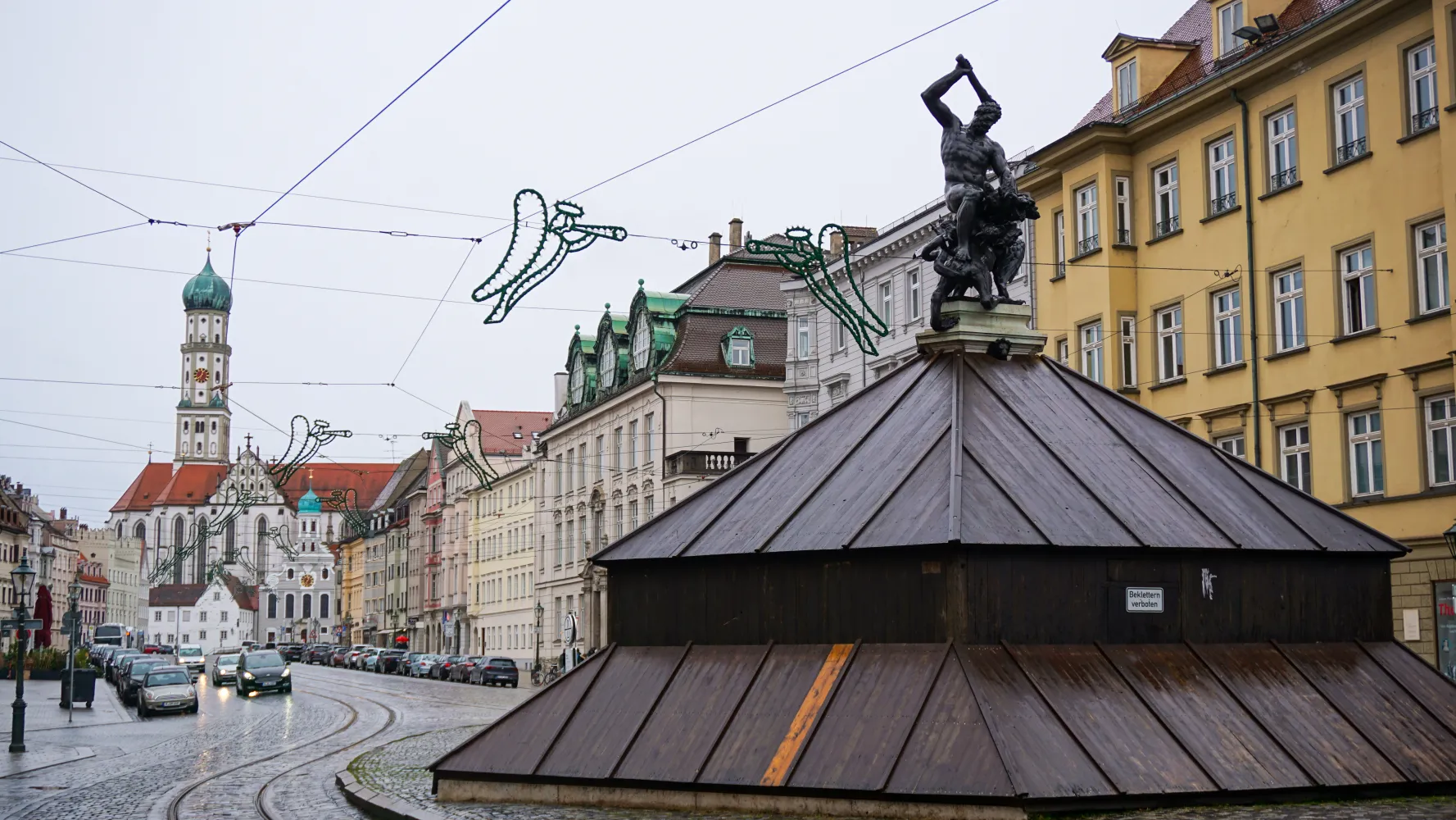 Street scene with a church tower, historic buildings, and a statue of a man wielding a hammer atop a wooden structure