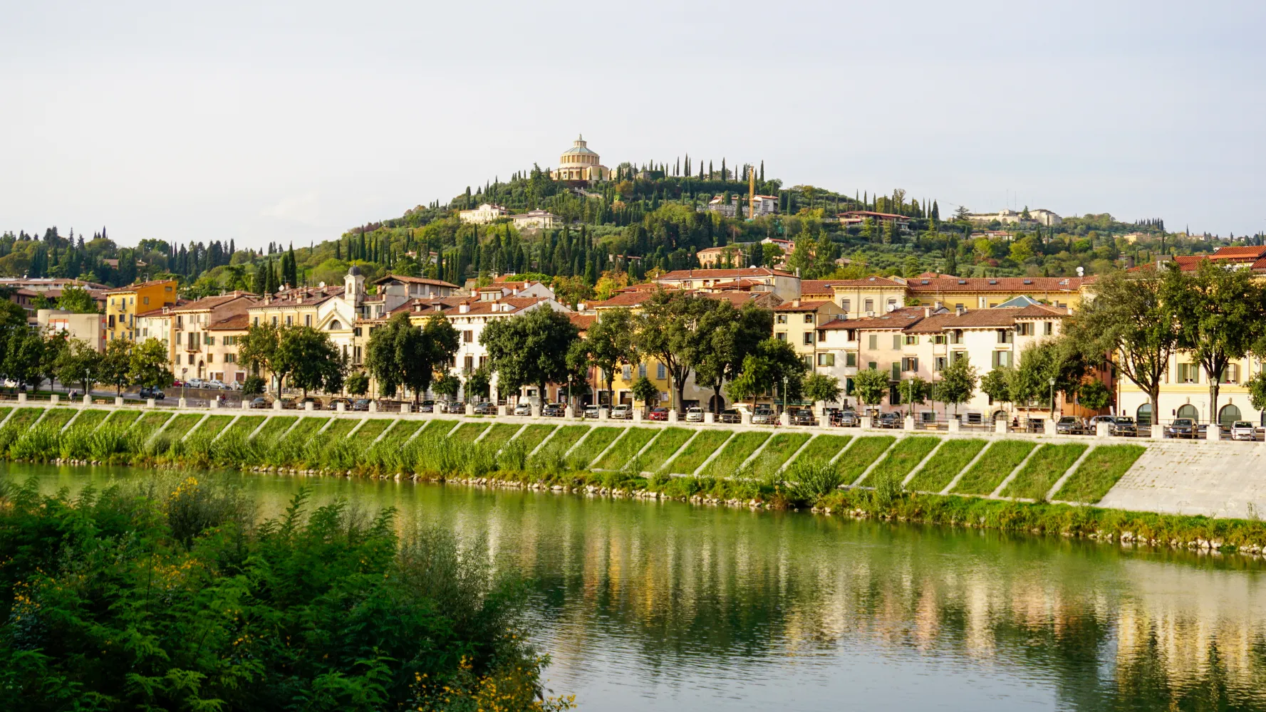 Riverfront with trees and buildings at the base of a green hill topped by a round structure
