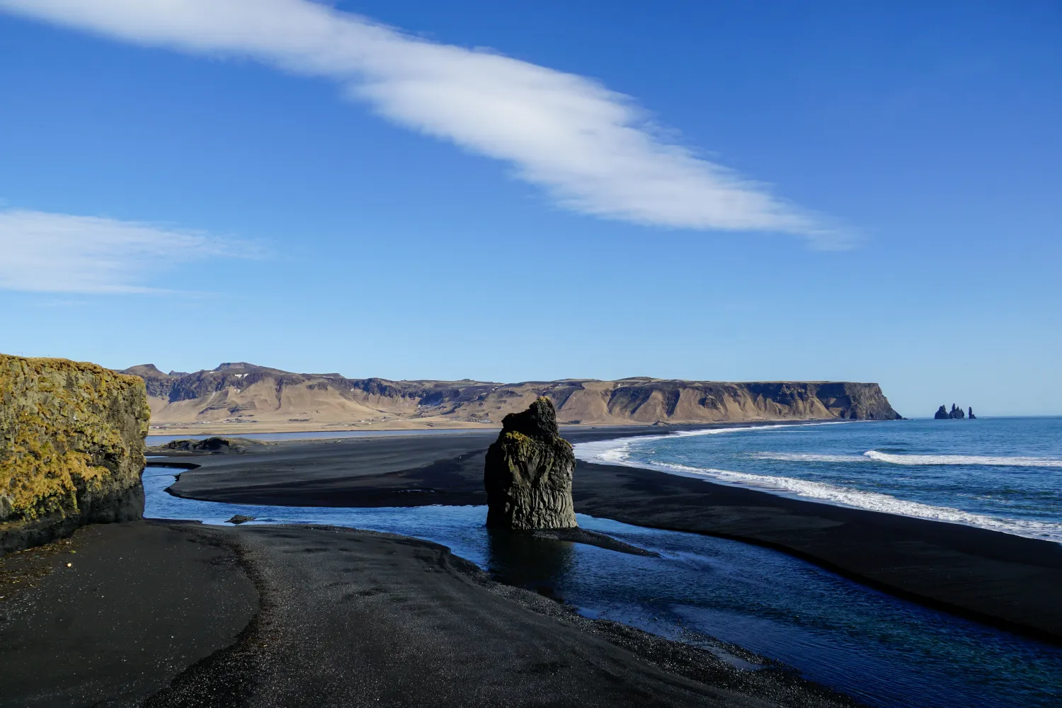 Black sand beach with a large rock formation, river, cliffs, and ocean under a blue sky